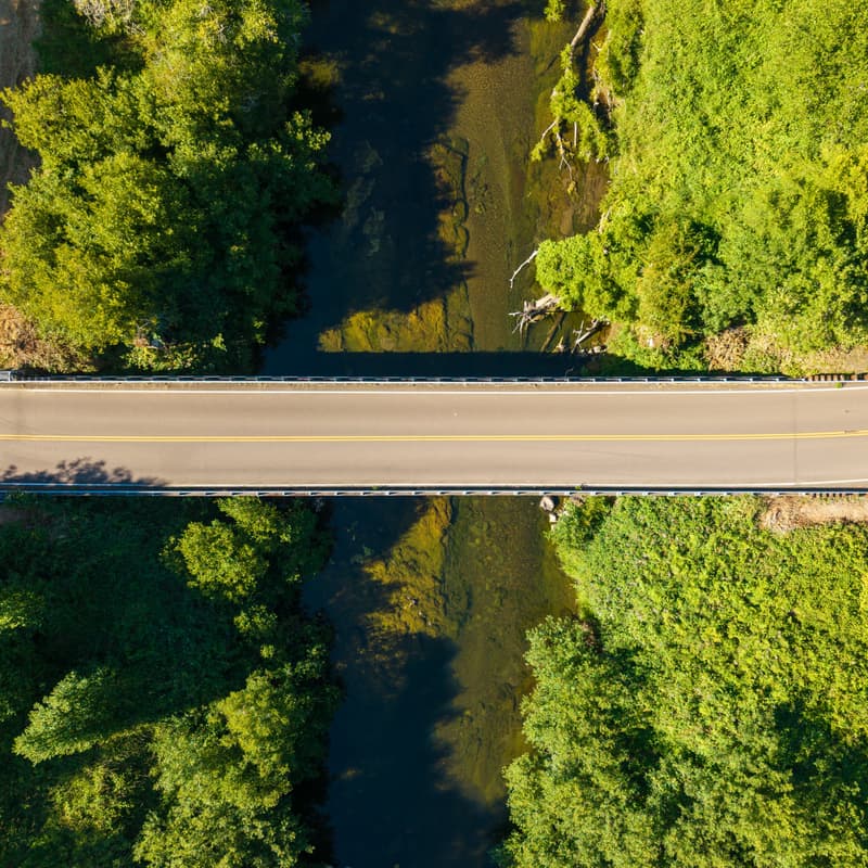 Top-down aerial of a two-lane road bridge crossing a dark river, framed by dense green trees on both banks