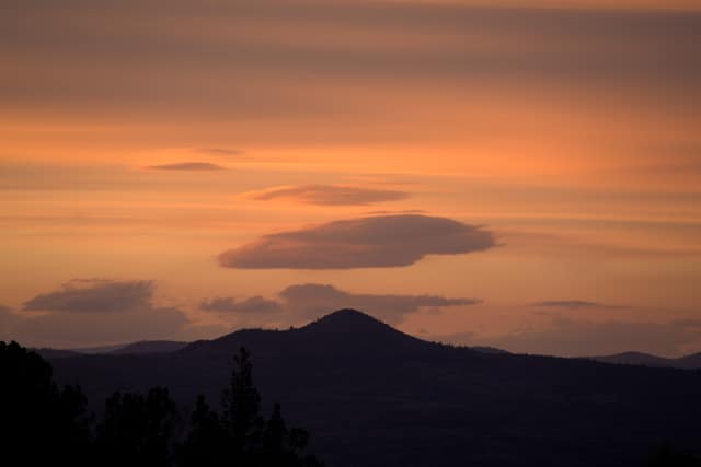 Lenticular clouds in an orange sunset sky over a dark butte